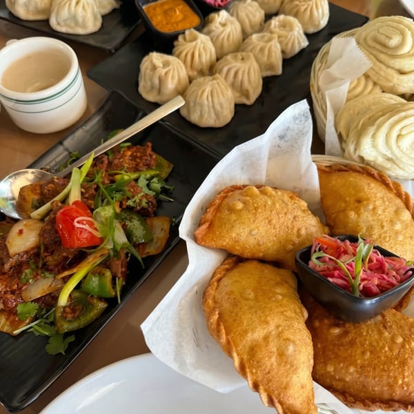 A table laden with various dishes, including momos, fried pastries, and a stir-fry, with dipping sauces.