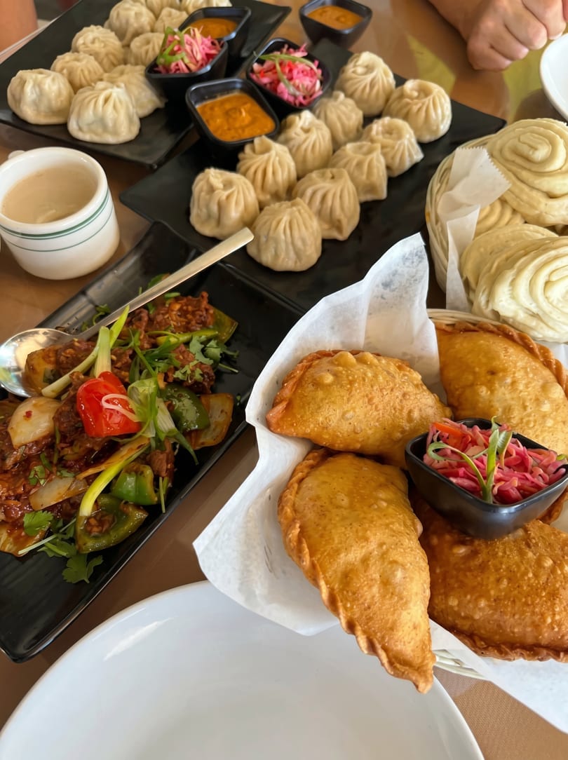 A table laden with various dishes, including momos, fried pastries, and a stir-fry, with dipping sauces.