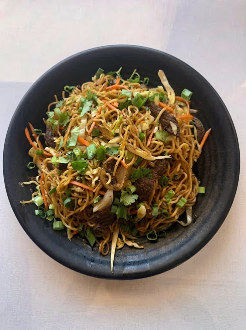 A black bowl filled with beef lo mein, garnished with green onions and cilantro.