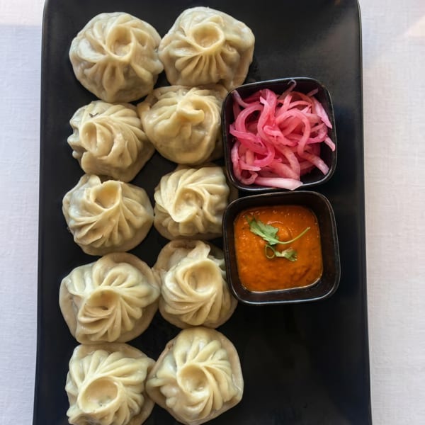 A black rectangular plate holds twelve steamed momos, red pickled onions, and orange chili sauce with cilantro.