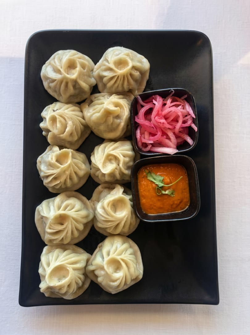 A black rectangular plate holds twelve steamed momos, red pickled onions, and orange chili sauce with cilantro.